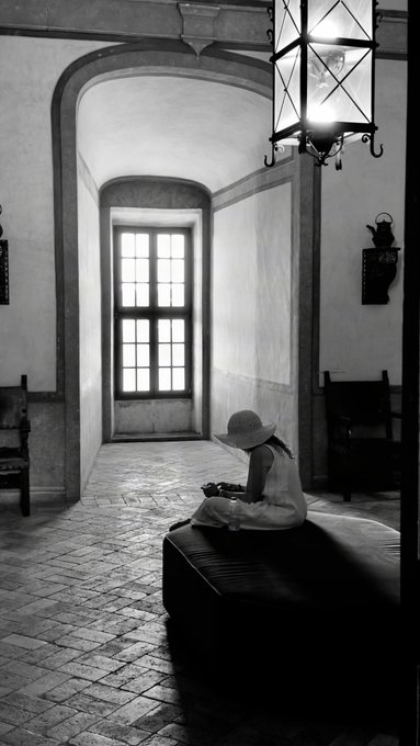 A black and white photograph of a child in a hat and dress sitting on a bench looking at an object, inside a stone hallway with a large window and a hanging lantern.