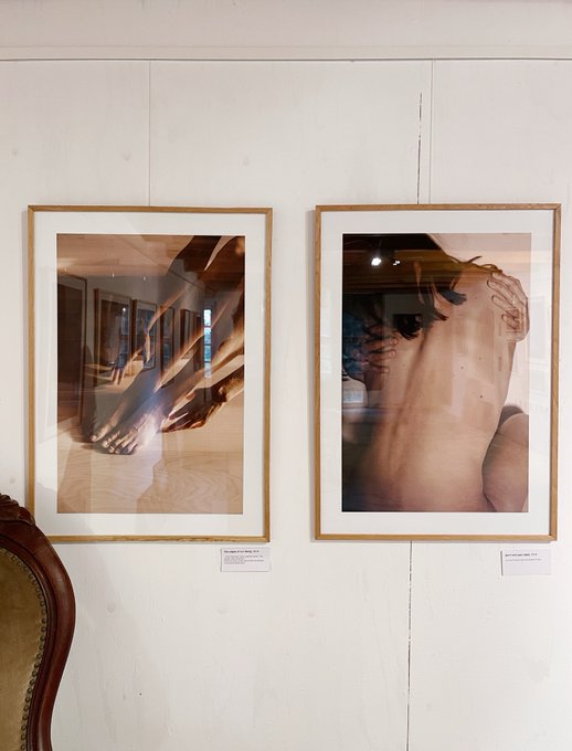 Two framed art photographs hang on a white paneled wall, depicting a pair of legs and a back, each with a small information placard below.