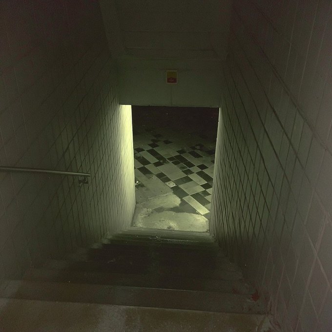 A dark, eerie view looking down a tiled stairwell into a hallway with a black and white checkered floor, appearing to be from a video game.