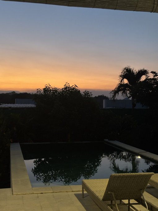A tranquil Bali sunset overlooks a private infinity pool, its still waters mirroring the warm orange sky and silhouettes of palm trees and tropical foliage, capturing the serene group retreat mentioned in the post.