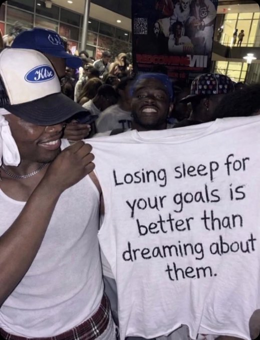 The image features two men standing next to each other with one of them holding up a t-shirt that says "Losing sleep for your goals is better than dreaming about them." The man wearing the hat has his hand over his mouth, possibly in surprise or shock at the message on the shirt. This scene takes place in front of a crowd, suggesting that they might be attending an event or gathering where this message was shared.