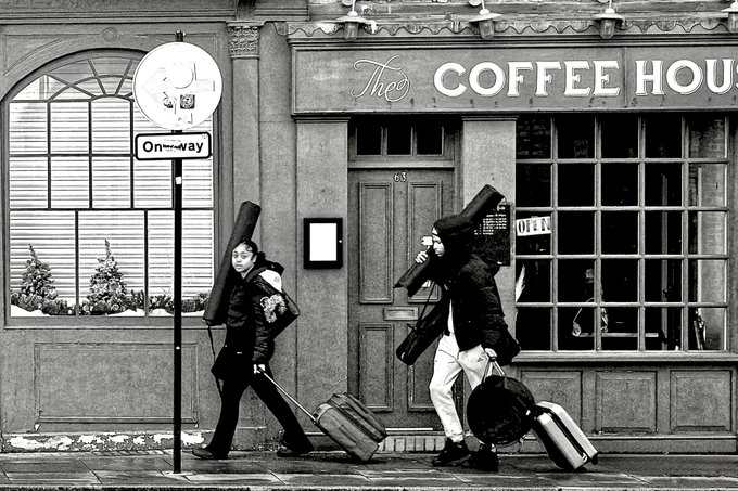 Two people walk along a city sidewalk carrying guitar cases and rolling suitcases past a storefront labeled "The COFFEE HOUSE," with a street sign beside them reading "One Way." The "One Way" sign ties into the caption's reference to "There's Only One Way 7," suggesting the image visually echoes the title's directional theme.