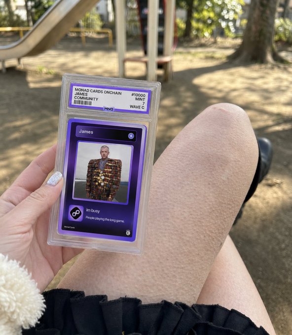 A hand with silver nail polish holds a graded 'Monad Cards Onchain' collectible card featuring a character named 'James,' with the player lounging outdoors near a playground slide.