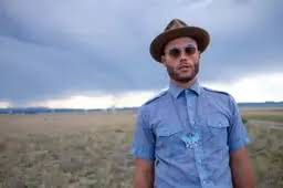 A man wearing a beige fedora, sunglasses, and a light blue button-up shirt with a turquoise necklace stands in a vast, grassy field under a dramatic, cloudy sky.