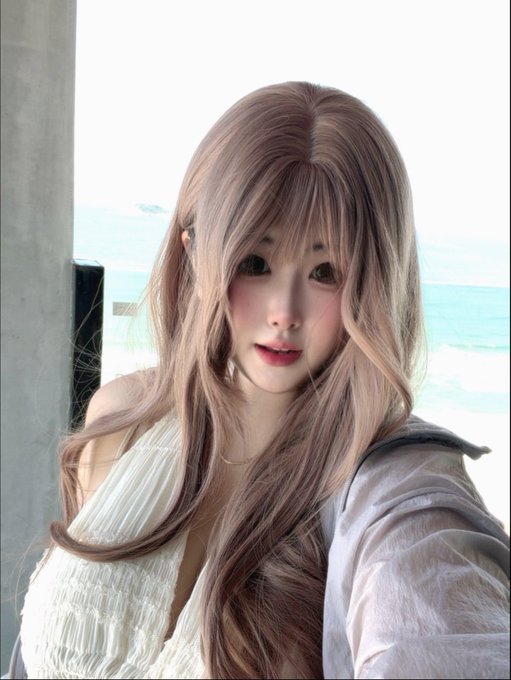 A young woman with long, flowing ash-blonde hair poses for a selfie by the sea, with turquoise ocean waters visible in the background. The image captures a serene seaside moment in Huizhou, embodying a peaceful "good morning" vibe along the coast.