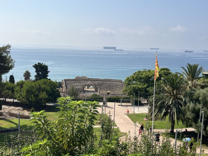 A scenic coastal view featuring ancient Roman amphitheater ruins in the foreground, with palm trees, a Mediterranean garden, and cargo ships visible on the sea.