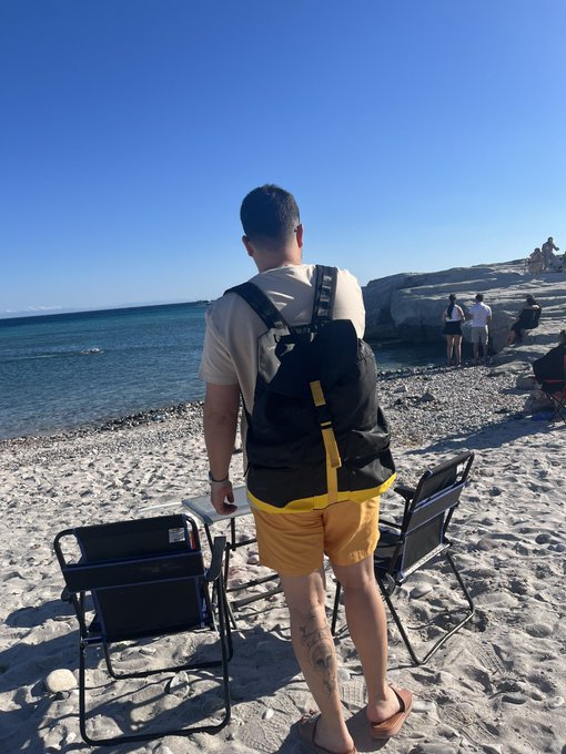 The image shows a man standing on a beach, wearing yellow shorts and carrying a black backpack with a yellow strap over his shoulder. There are two chairs visible near him, one of which has a blue cover. The background features an ocean view under a clear sky.