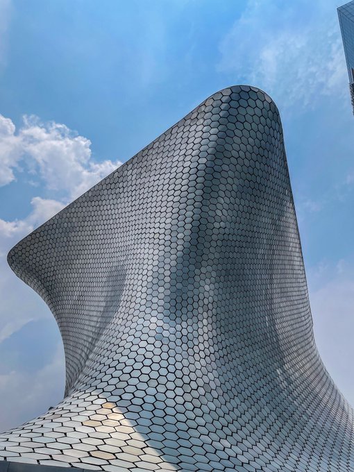 A modern, curved building facade covered in dark hexagonal tiles, photographed from below against a blue sky with scattered clouds.