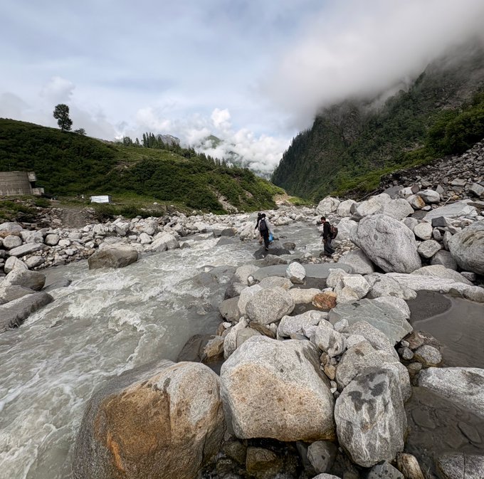 A scenic landscape of a rushing mountain river flowing over large gray boulders, with two people crossing the water; misty green mountains and a cloudy sky fill the background.