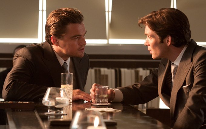 A cinematic still of two men in suits (Leonardo DiCaprio and Cillian Murphy) sitting at a bar, staring intensely at each other.