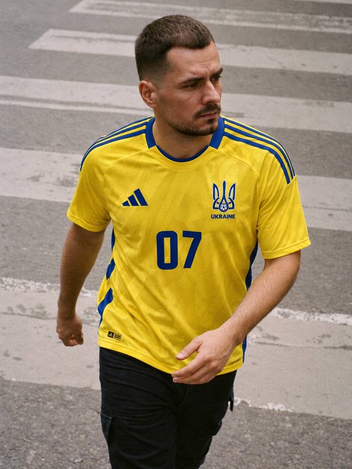 A man with short hair and a mustache wearing a yellow Ukraine national team jersey with number 07 and the Adidas logo walks across a crosswalk.