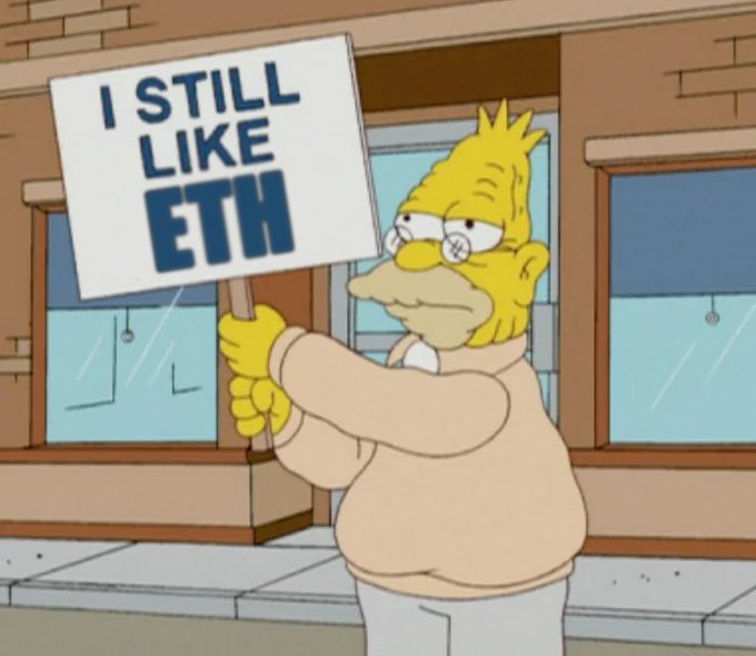 The image features a cartoon character, likely Homer Simpson from the Simpsons TV show, holding up a sign that reads "I Still LikeETHing". The character appears to be standing outside of a building or store, possibly a clothing shop as suggested by the context provided in the caption. The scene is set against an urban backdrop with buildings and windows visible in the image.