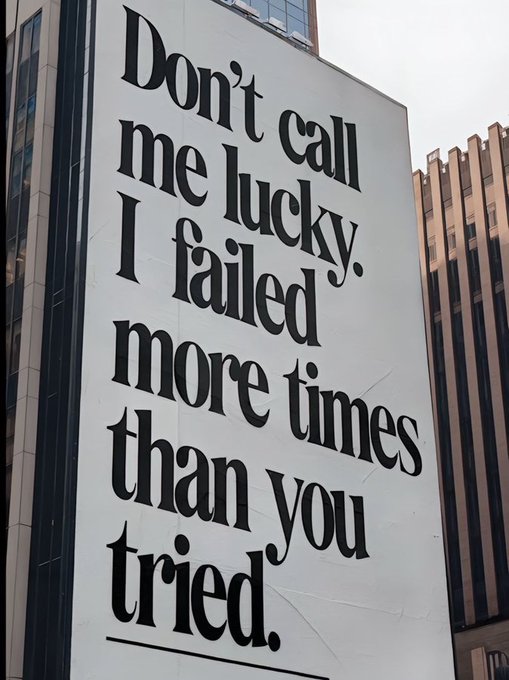 A large building billboard reads, “Don’t call me lucky. I failed more times than you tried.” The caption challenges viewers to understand that persistence through repeated failure—not luck—is the true source of success.