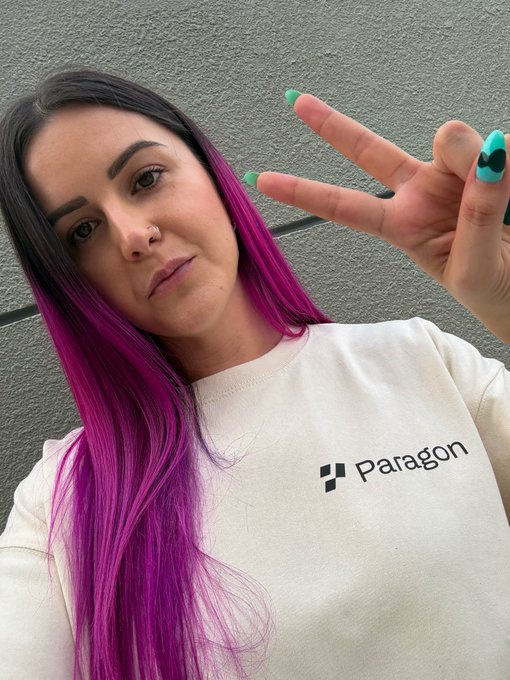 A woman with long purple and dark hair takes a selfie while making a peace sign, wearing a sweatshirt branded with the 'Paragon' logo.