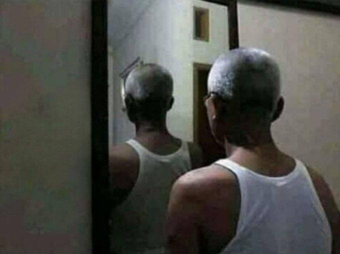 A photograph showing an older man in a white tank top standing in front of a mirror, looking at his own reflection.