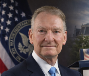 The photo appears to be an official portrait of a man in a dark suit and blue tie, posed in front of an American flag and what looks like a U.S. government seal (likely the Treasury or SEC emblem), suggesting a senior U.S. official. The caption accompanying the post about the SEC's canceled crypto meeting likely identifies him as the SEC official responsible for the delayed "Clarity" crypto regulation announcement.