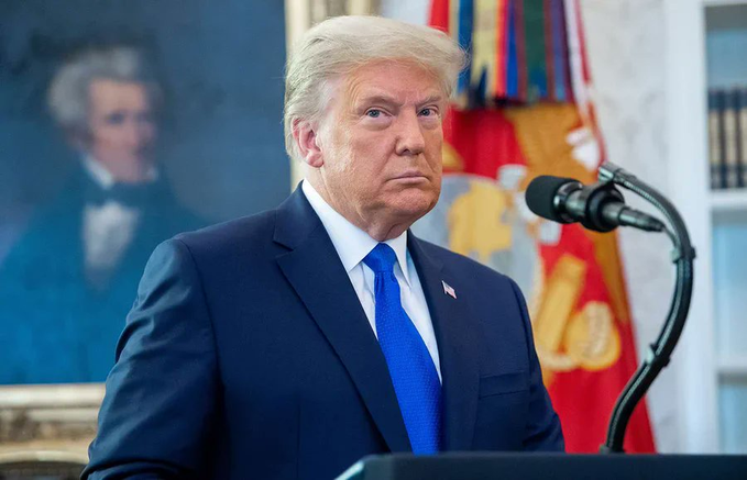 Donald Trump, wearing a dark blue suit and bright blue tie, stands at a podium in front of microphones with a portrait painting and military flags visible behind him.
