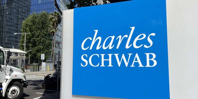 The image shows a blue sign with white text that reads "Charles Schwab" against a gray background. The sign is located near a street where a truck can be seen parked next to it. In the background, there are tall buildings visible behind the truck.