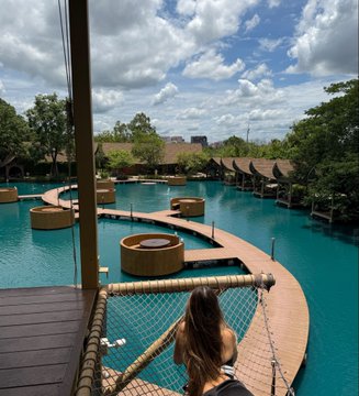 A person with long brown hair stands on a wooden deck overlooking a winding turquoise pool complex lined with circular platforms and tropical trees, evoking a "Solana summer" vibe of leisure and warmth. The bright, sunny scene with clear skies and lush greenery captures the relaxed, resort-like atmosphere the caption suggests.