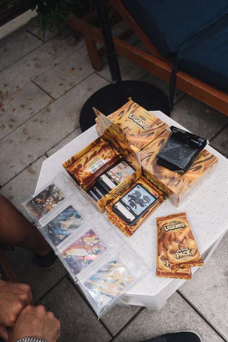 A collection of Pokémon TCG products, including a booster box, packs, and cards in protective sleeves, is laid out on a small white table outdoors.