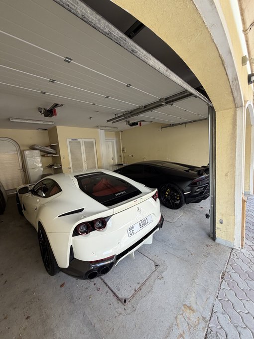 A white Ferrari and a black Lamborghini are parked in a garage. The Ferrari's license plate reads "CB K8077," indicating it's likely registered in the Bahamas. Given the context of "GM," both cars are iconic European exotic brands, contrasting with American General Motors vehicles.