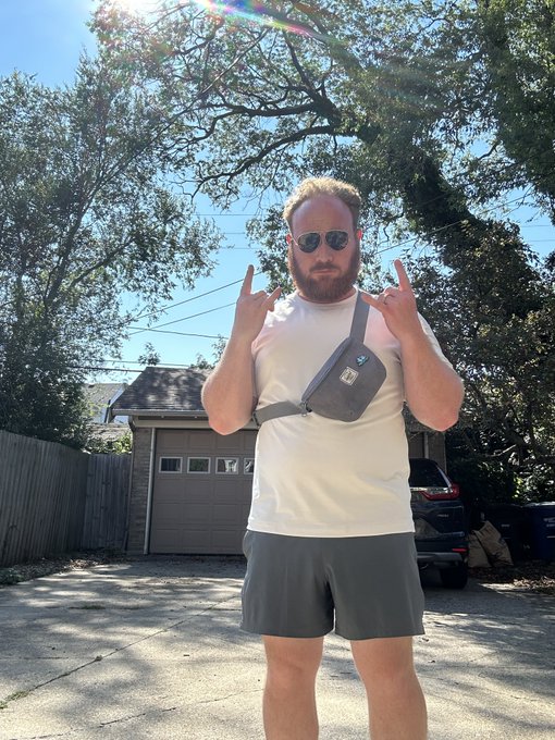 A man with a red beard and sunglasses stands in a driveway wearing a white t-shirt, gray shorts, and a black crossbody bag, making hand gestures under sunny skies.