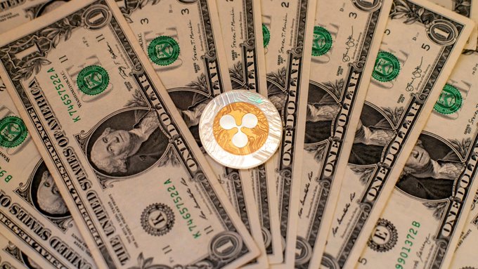 A fanned-out spread of US one-dollar bills topped with a physical silver and gold Ripple (XRP) cryptocurrency coin.