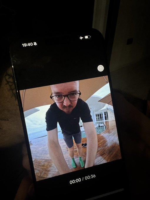 The image shows a man wearing glasses, standing under an umbrella indoors with his phone held up next to him. There are two other people visible in the background, but they are not the main focus of the photo. The text "SOLANA SUMMER BTS" is also present in the image.