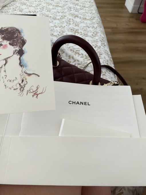 The image shows a burgundy quilted Chanel handbag with gold hardware, displayed alongside Chanel-branded white packaging and a card featuring a watercolor sketch of a woman. The "CHANEL" text is prominently visible on the packaging, suggesting a luxury shopping or unboxing moment.