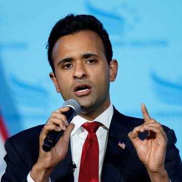 A man in a dark suit and red tie speaks into a microphone while gesturing with his hand, with an American flag pin on his lapel.