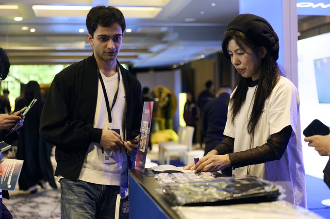 Two young people, a man in a black jacket and a woman wearing a black beret, stand at a booth interacting with merchandise at what appears to be a tech or crypto conference.