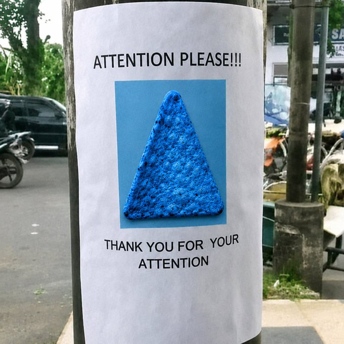 A printed poster taped to a pole that reads "ATTENTION PLEASE!!!" above an image of a blue, textured triangular object resembling a Dorito, and "THANK YOU FOR YOUR ATTENTION" below it.