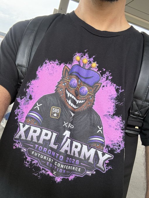 A person wearing a black t-shirt featuring an XRPL Army graphic of a grinning cat in a purple beret and sunglasses, with text reading "XRPL ARMY, TORONTO 2026, FUTURIST CONFERENCE EDITION," along with a black backpack, perfectly matching their caption about attending Canada's largest Web3 & Crypto conference as an XRPL supporter.
