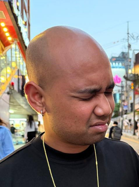 A close-up selfie of a young bald man with a silver hoop earring and gold chain, making a displeased facial expression with closed eyes in an urban street setting with neon signs.