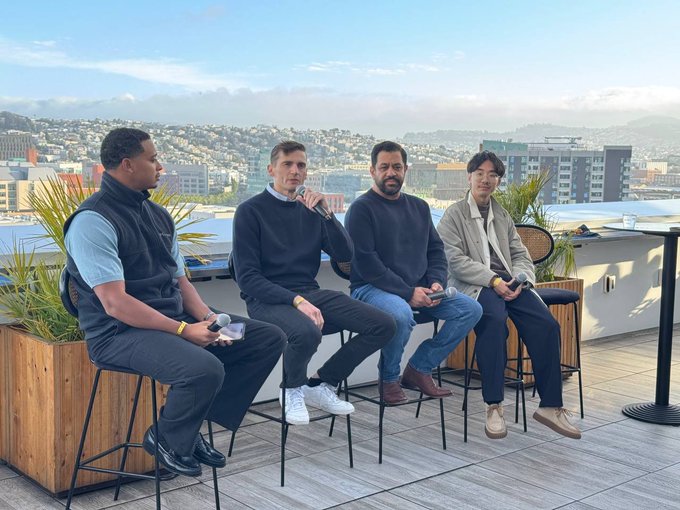 The image shows four men sitting at a table with their legs crossed, each holding a remote control. They are all wearing black clothing and appear to be enjoying themselves as they watch television or engage with some other form of media. The setting appears to be an outdoor patio area, possibly on a rooftop, which adds to the relaxed atmosphere. The image does not provide any specific information about the content being watched or the event that took place during their gathering.