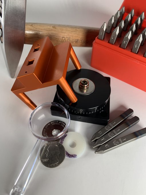 A photograph of metalworking tools including a hammer, an orange anvil-shaped tool, a hammer head, screwdrivers, engraving bits, a magnifying glass, and a coin on a white surface.