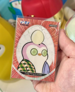 A hand holds up a Pokémon trading card sleeve featuring "Pudgy Penguin #1589". In the blurred background, several colorful plush toys, including what appears to be a yellow Pokémon, are visible.