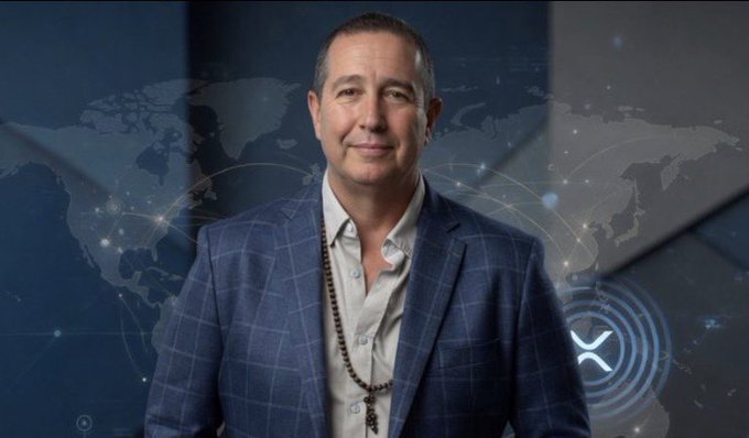The image features a man wearing a blue suit standing against a blue background with a map of the world behind him, as well as an XRP symbol in the bottom right corner. The caption reads "Just In: @DigPerspectives says 'The Stage Is Set For $XRP Supply Shock.'". This suggests that there is a significant development or event related to cryptocurrency and its supply chain, which has caught the attention of the man wearing the suit.