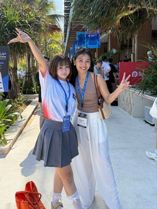 Two young women pose for a photo at what appears to be a convention or event, both wearing lanyards with badges. They are smiling and making peace signs, with tropical plants visible in the background.