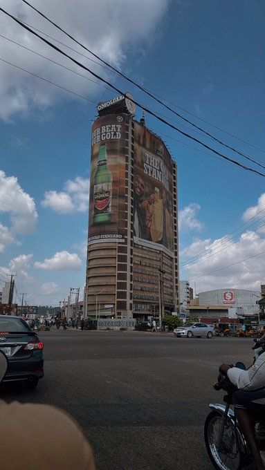 A tall circular building features a large Guinness advertisement reading "GET BEAT FOR GOLD" alongside "THE STAND" billboard, while traffic moves through a busy intersection below under a partly cloudy sky—a bustling urban scene that captures the weariness of navigating a hectic day.