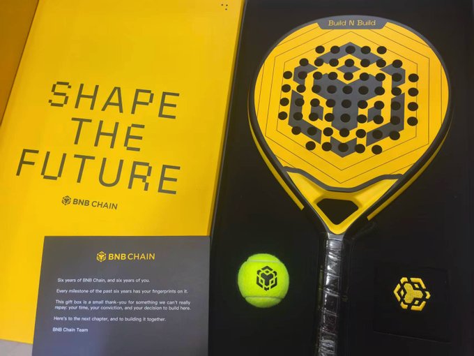A BNB Chain promotional display featuring a yellow “SHAPE THE FUTURE” panel, a BNB-branded padel racket, and ball, plus a “Build N Build” label and the slogan “Here’s to the next chapter, and to building it together.”