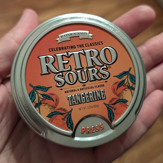 A hand holds a circular tin of "Retro Sours" tangerine-flavored candy, labeled "Iconic Candy," "Celebrating the Classics," and "Press to open," with vintage-style citrus illustrations on the label—reflecting the post's excitement about the beloved candy making a nostalgic comeback.