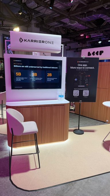 A Karrier One exhibition booth in Singapore features a large display stating “Billion are still underserved by traditional telecom,” with statistics of 5B, 3B, and 2B, while a neighboring sign reads “One app. More ways to connect” and invites visitors to scan a QR code. The setup includes the illuminated Karrier One sign, a presentation screen, a counter, a water bottle, and a pink-and-white chair, matching the post’s invitation to visit the booth and try the new Karrier One app.