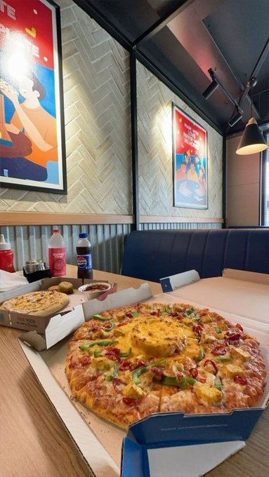 Pizza Hut restaurant interior with a pineapple-topped pizza in the foreground, condiment bottles (ketchup, chili sauce), garlic bread, and wall art posters.