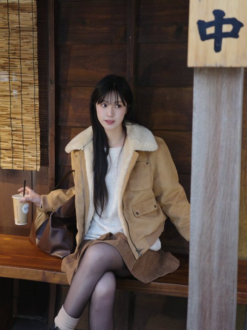 A young woman in a tan shearling jacket sits on a wooden bench holding an iced coffee, with a Japanese sign reading "中" visible beside her. The image contrasts the calm, composed scene with the volatile GRASS cryptocurrency price action described in the post, where the coin briefly broke past $0.8 before pulling back.