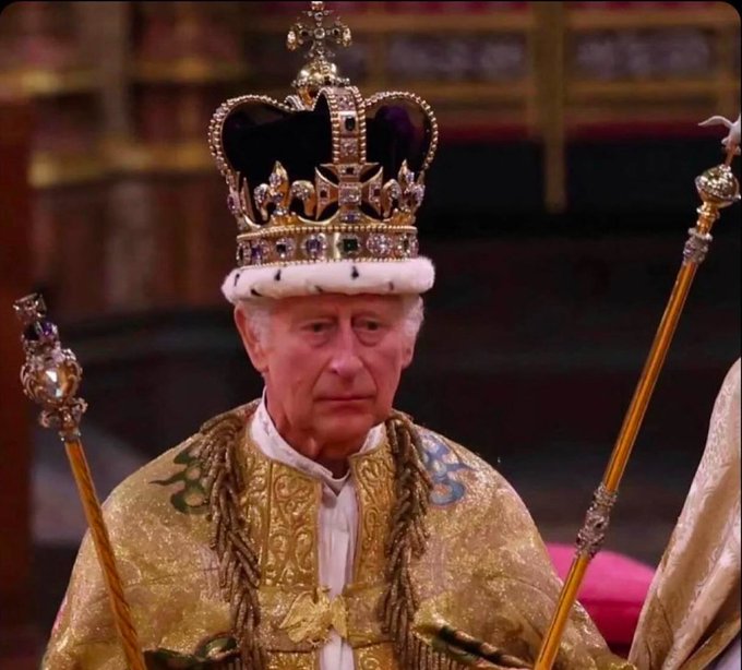 King Charles III wearing a crown and golden ceremonial robes, holding two ceremonial scepters.
