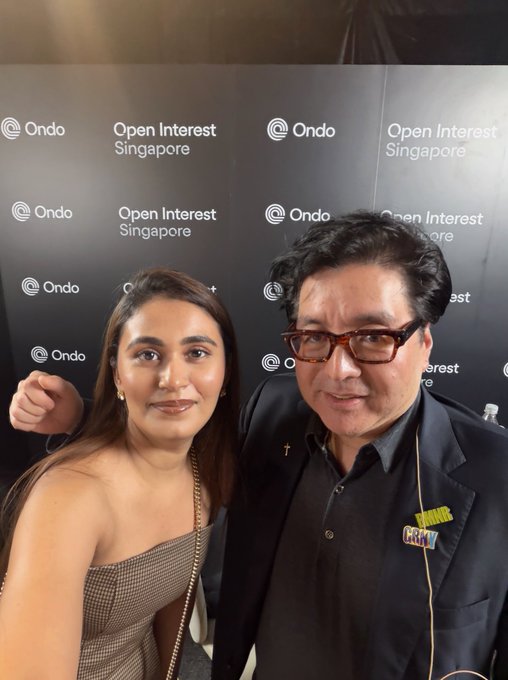 A man and woman pose for a selfie in front of a black backdrop featuring "Ondo" and "Open Interest Singapore" logos, kicking off Token2049 in style.