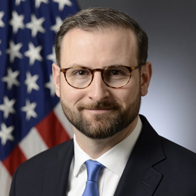 White House crypto adviser Patrick Witt is pictured here, smiling and wearing glasses with a blue tie, posed in front of an American flag in an official portrait setting. The image accompanies news of his remarks about feeling "very good" ahead of a Senate procedural vote on a bipartisan crypto bill, highlighting his role in advocating for the legislation's passage.