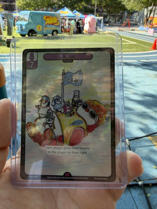 A hand holding a holographic 'Selfish' action card (cost 7) featuring illustrated penguins in a car, with a game tent visible in the background.