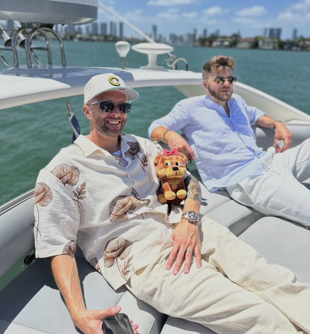 Two men relax on a yacht; one in the foreground wears a 'C' cap and holds a small stuffed lion toy, while the other wears sunglasses and a blue striped shirt.
